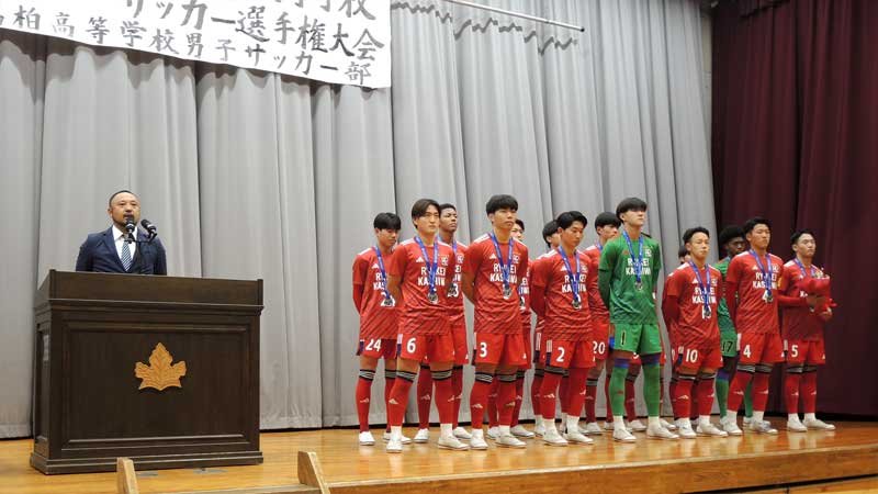 流通経大柏高校で開かれた準優勝報告会でサッカー部員らと並びあいさつする榎本監督＝１５日、柏市