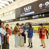 成田空港からのタクシー利用を促す広告を表示した国際線到着ロビーで、案内小冊子を手渡す空港スタッフら＝同日、成田市